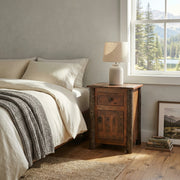 Horseshoe Mountain Barnwood and Hickory Enclosed Nightstand with a lamp beside a bed with a striped throw near a window