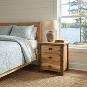 Mount Flora Hickory Log Three Drawer Nightstand in Natural with a lamp beside a bed with blue bedding near a lake-view window